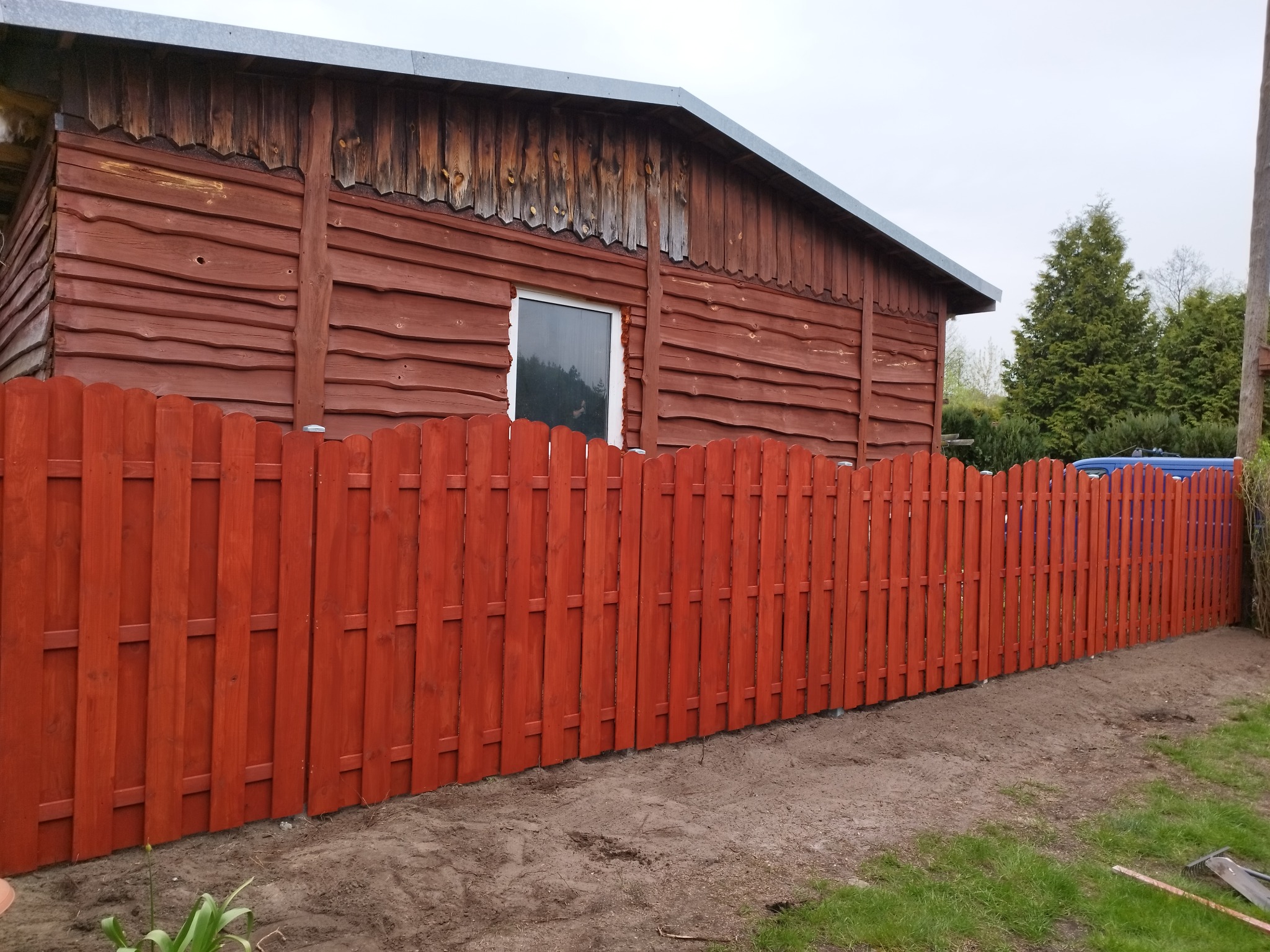 Czerwony drewniany płot sztachetowy przed drewnianym budynkiem z szarym dachem, widok z boku na nierówny teren.