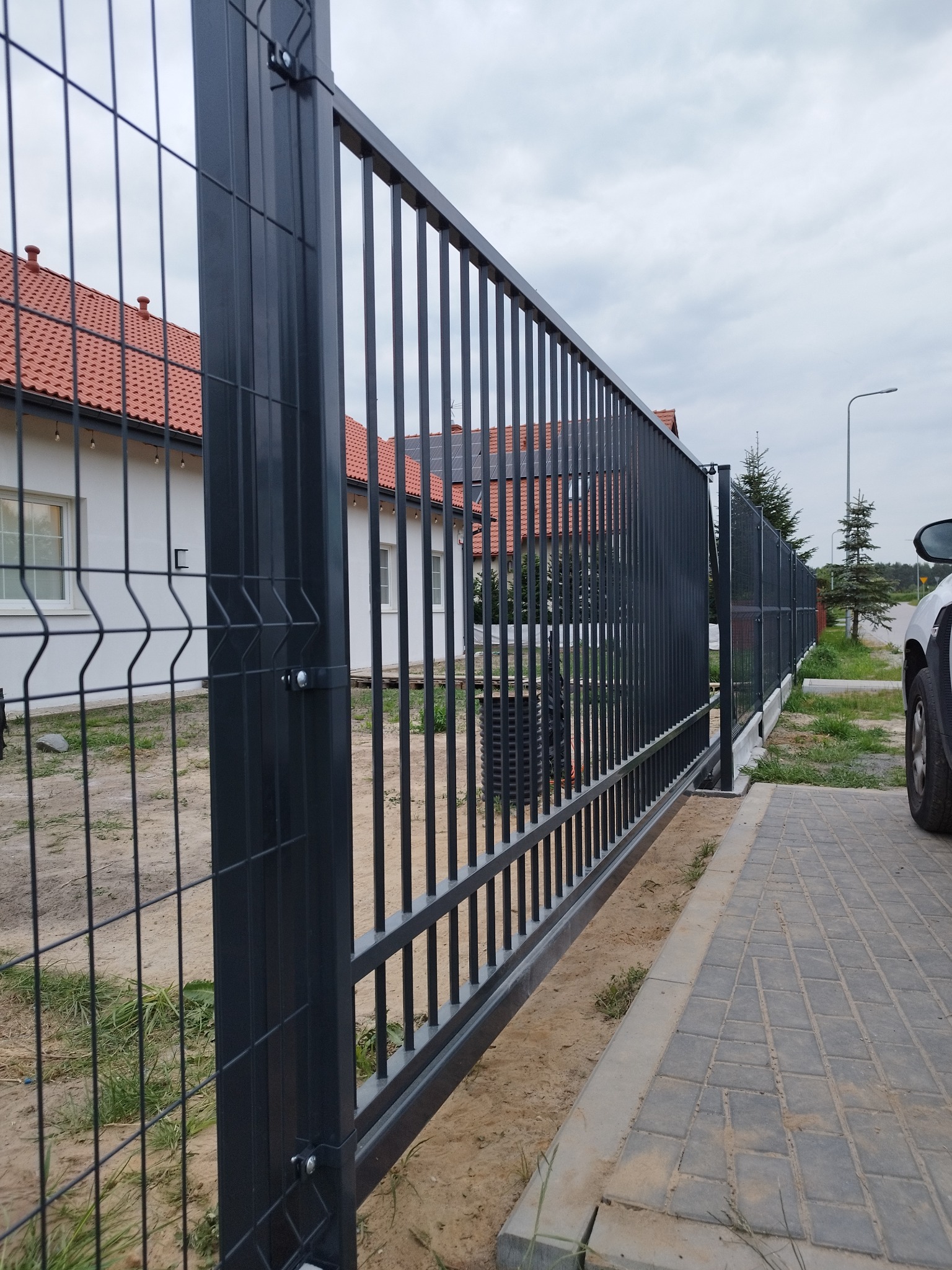 Nowoczesne, grafitowe ogrodzenie panelowe z furtką, widok z boku, kostka brukowa, dom z czerwonym dachem w tle.