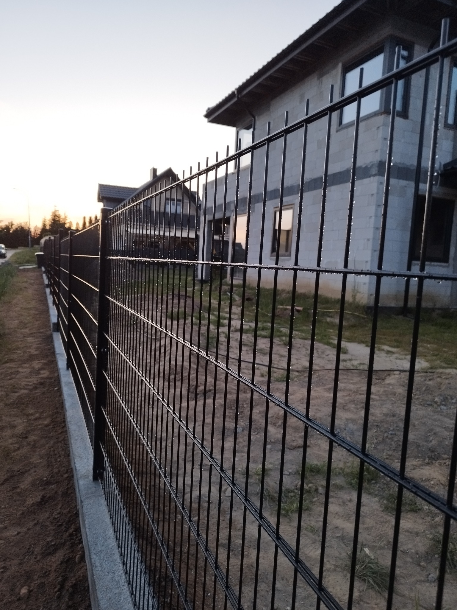 Nowoczesne czarne ogrodzenie panelowe z betonu, widoczne krople wody, w tle surowy budynek w trakcie budowy i zachodzące słońce.