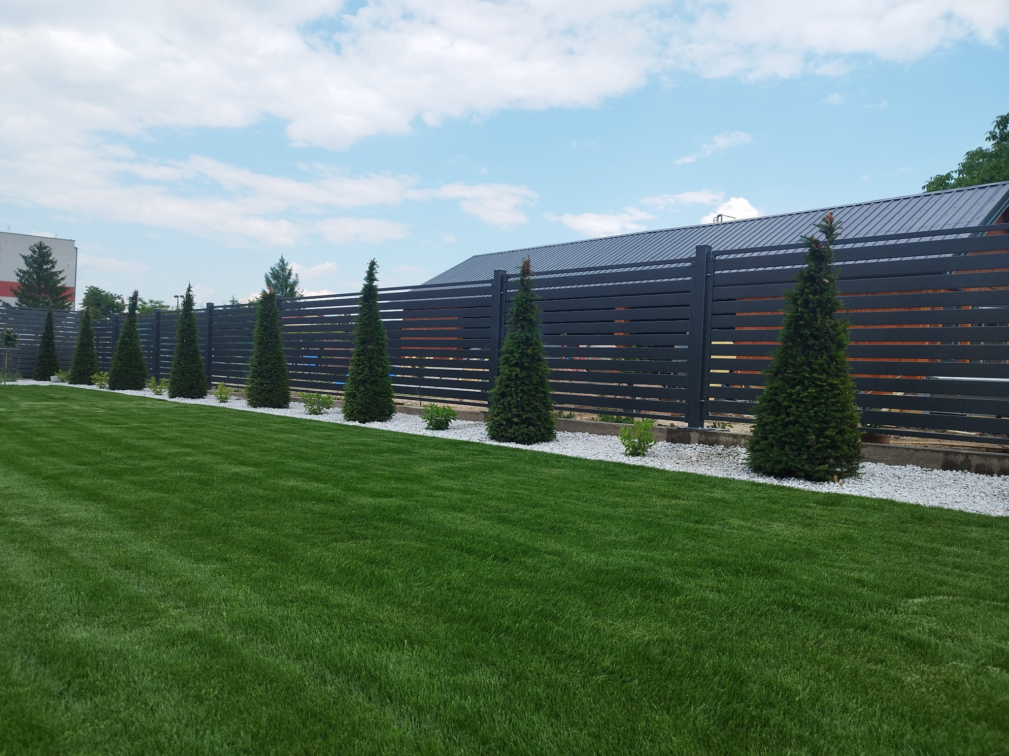 Nowoczesne ogrodzenie panelowe w kolorze grafitowym, z poziomymi szczeblami, oddzielające wypielęgnowany trawnik od posesji, obsadzone tujami i drobnymi krzewami na tle białego grysu