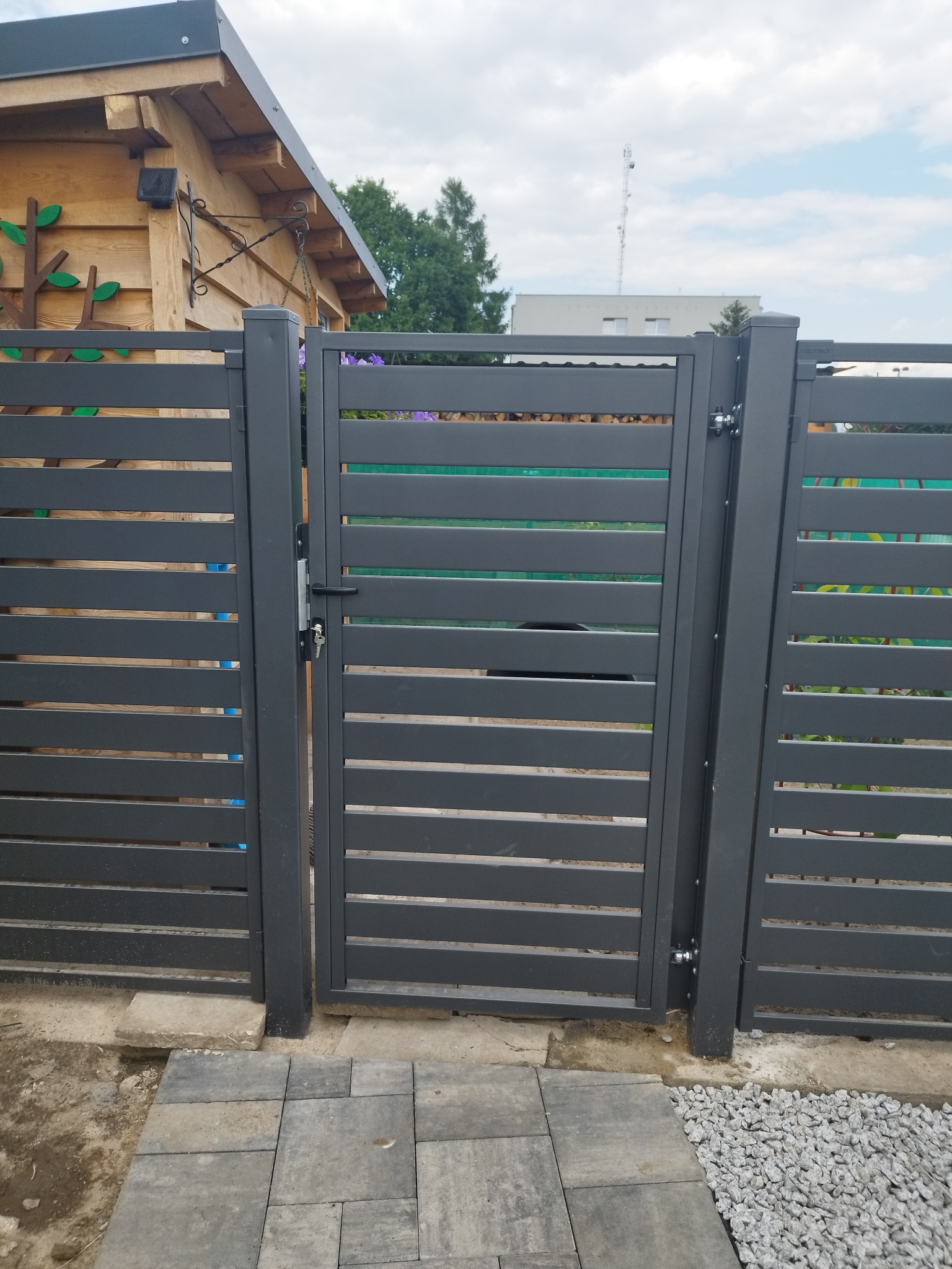 Nowoczesna, szara furtka panelowa z poziomymi szczeblami, z widocznym zamkiem i kluczem, prowadząca do ogrodu z drewnianą altaną i fragmentem kostki brukowej.