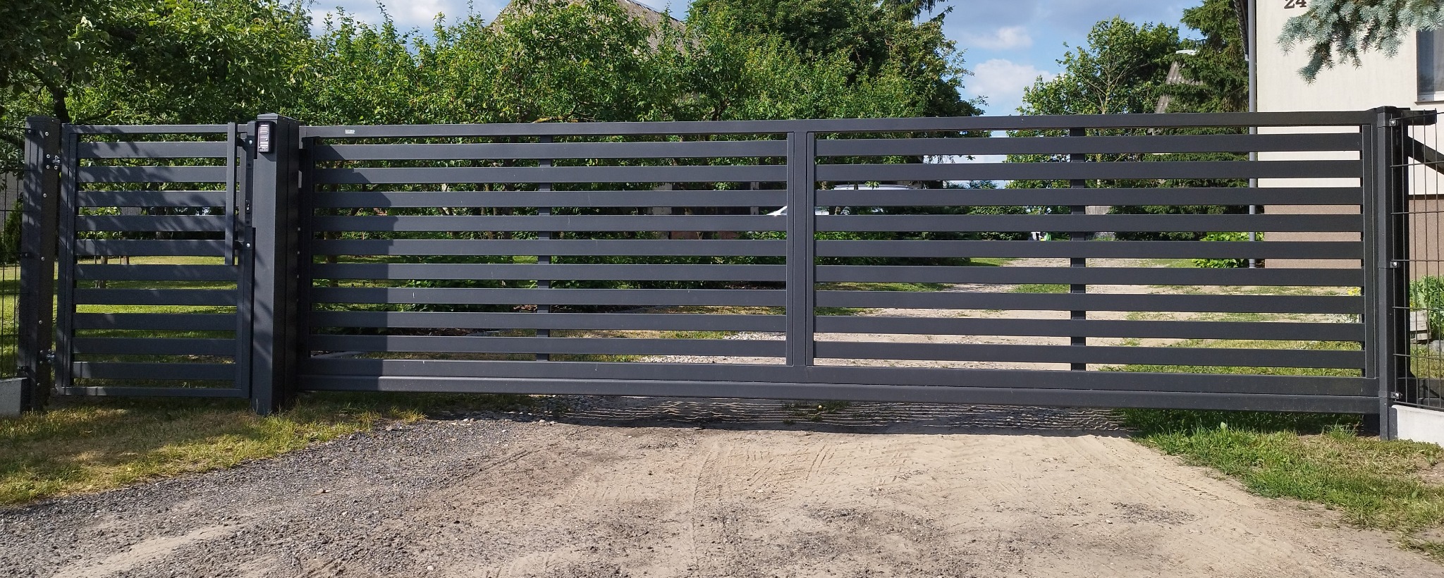 Nowoczesna, przesuwna brama w kolorze grafitowym z poziomymi panelami, zamontowana na posesji z widocznym numerem domu i zielenią w tle.