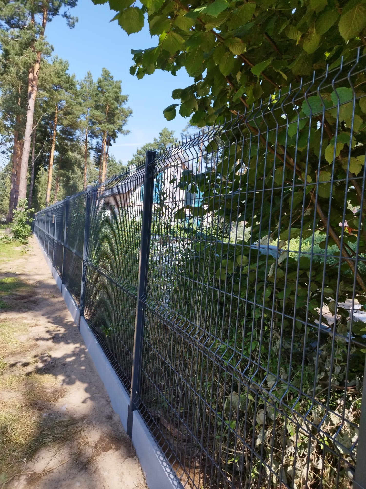 Długie, panelowe ogrodzenie w kolorze grafitowym, zamontowane na betonowej podmurówce, otoczone zielenią drzew i krzewów, z widocznym budynkiem w tle.