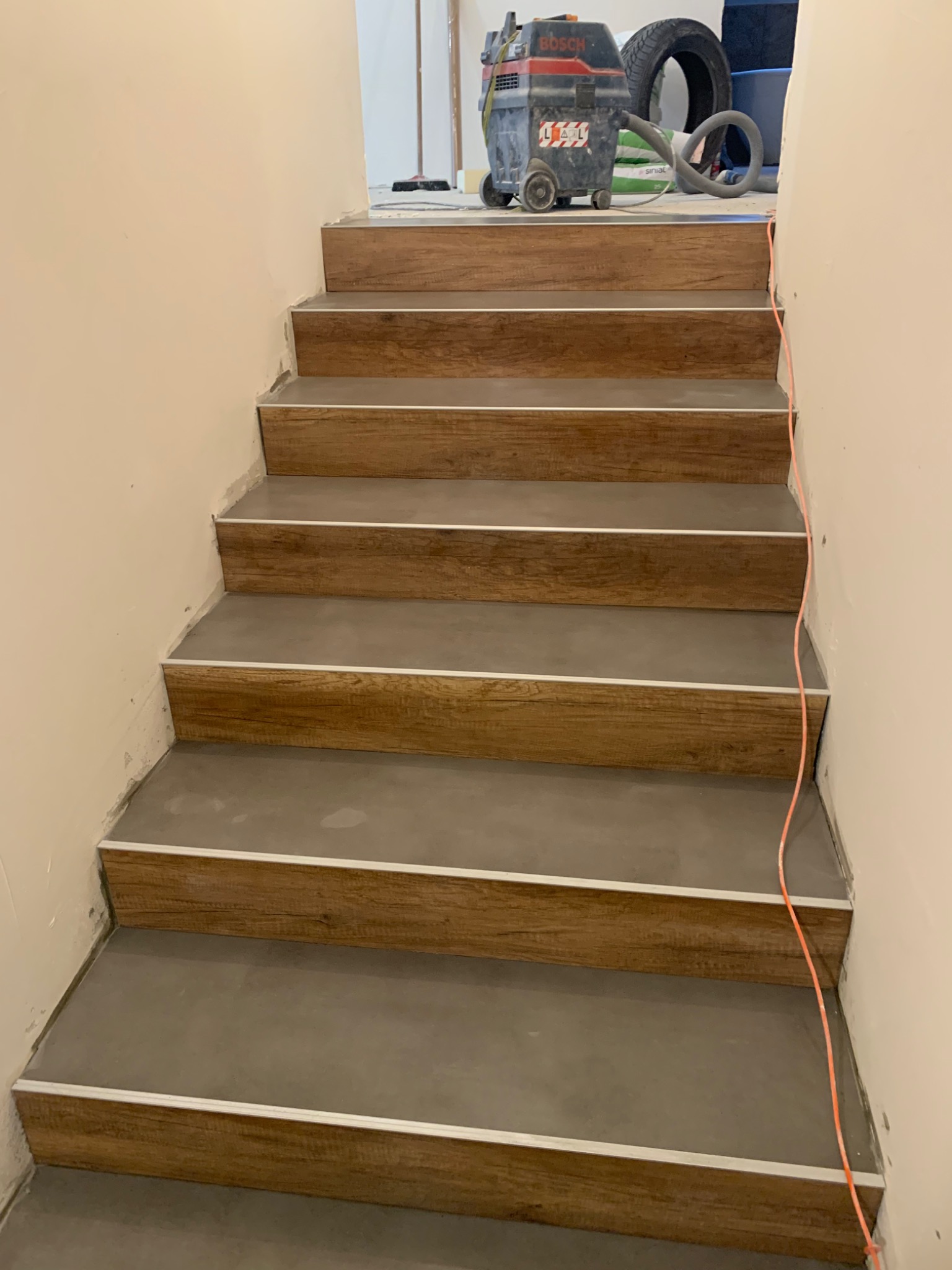 Schody wykończone panelami imitującymi drewno, z szarymi stopnicami i srebrnymi listwami, w tle odkurzacz przemysłowy Bosch i opona samochodowa.