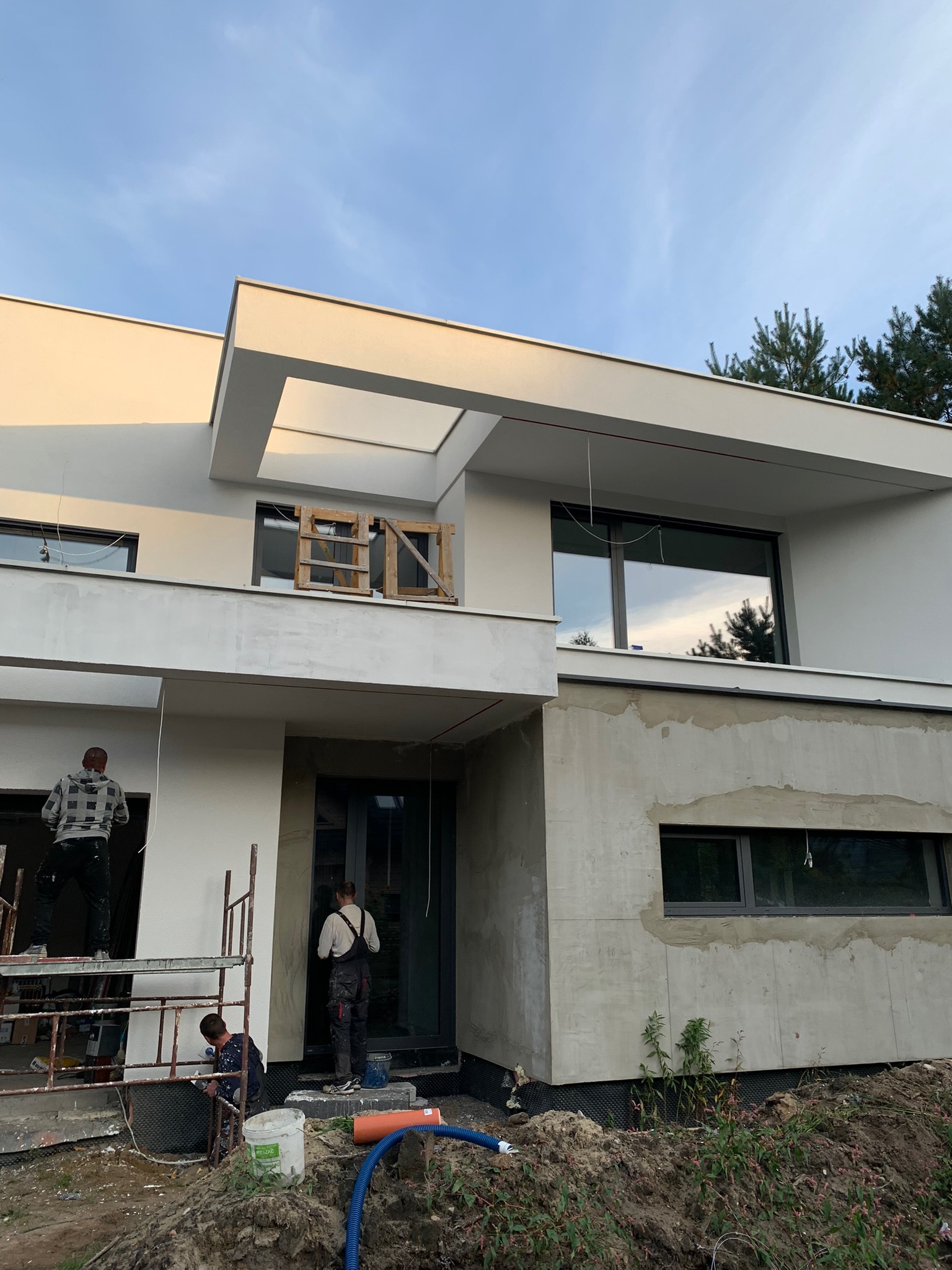 Ocieplanie ścian zewnętrznych nowoczesnego domu jednorodzinnego, widoczni pracownicy na rusztowaniu i przy wejściu, część elewacji w surowym tynku.