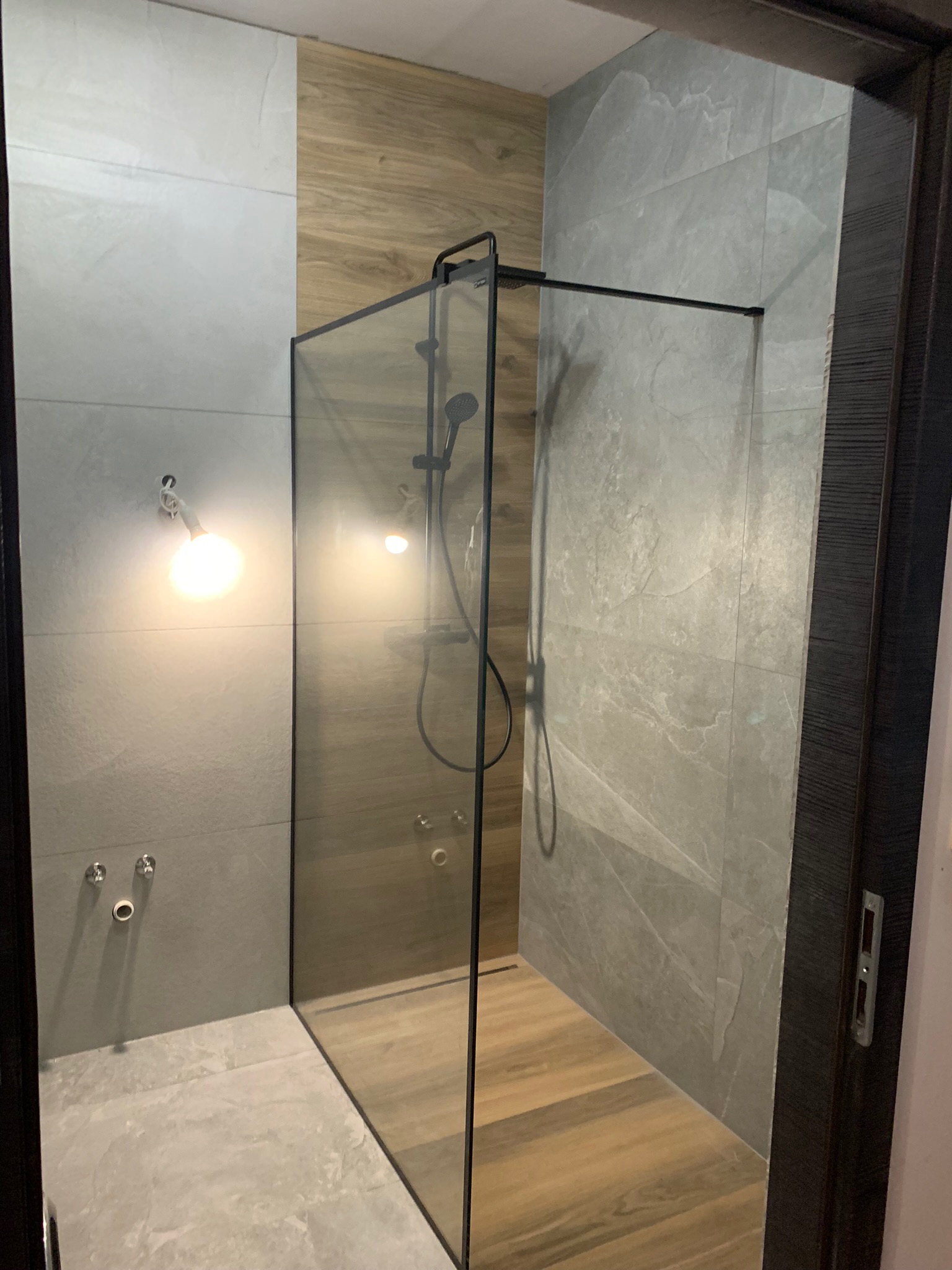 Kabina prysznicowa typu walk-in z czarnymi profilami, obłożona płytkami imitującymi drewno i szary kamień, oświetlona punktowym światłem.