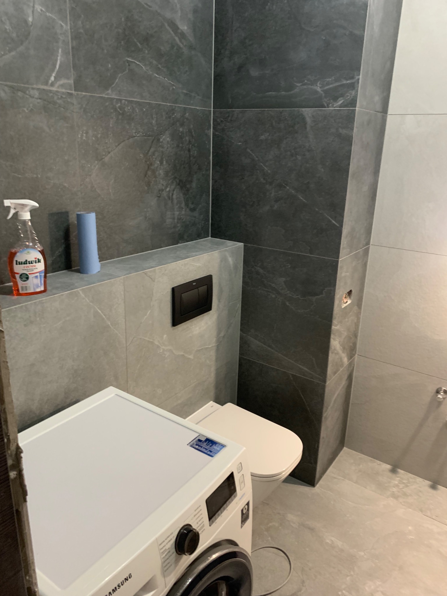 Nowoczesna łazienka z szarymi płytkami imitującymi kamień, białą toaletą i pralką Samsung, na półce środki czystości Ludwik i rolka papieru.
