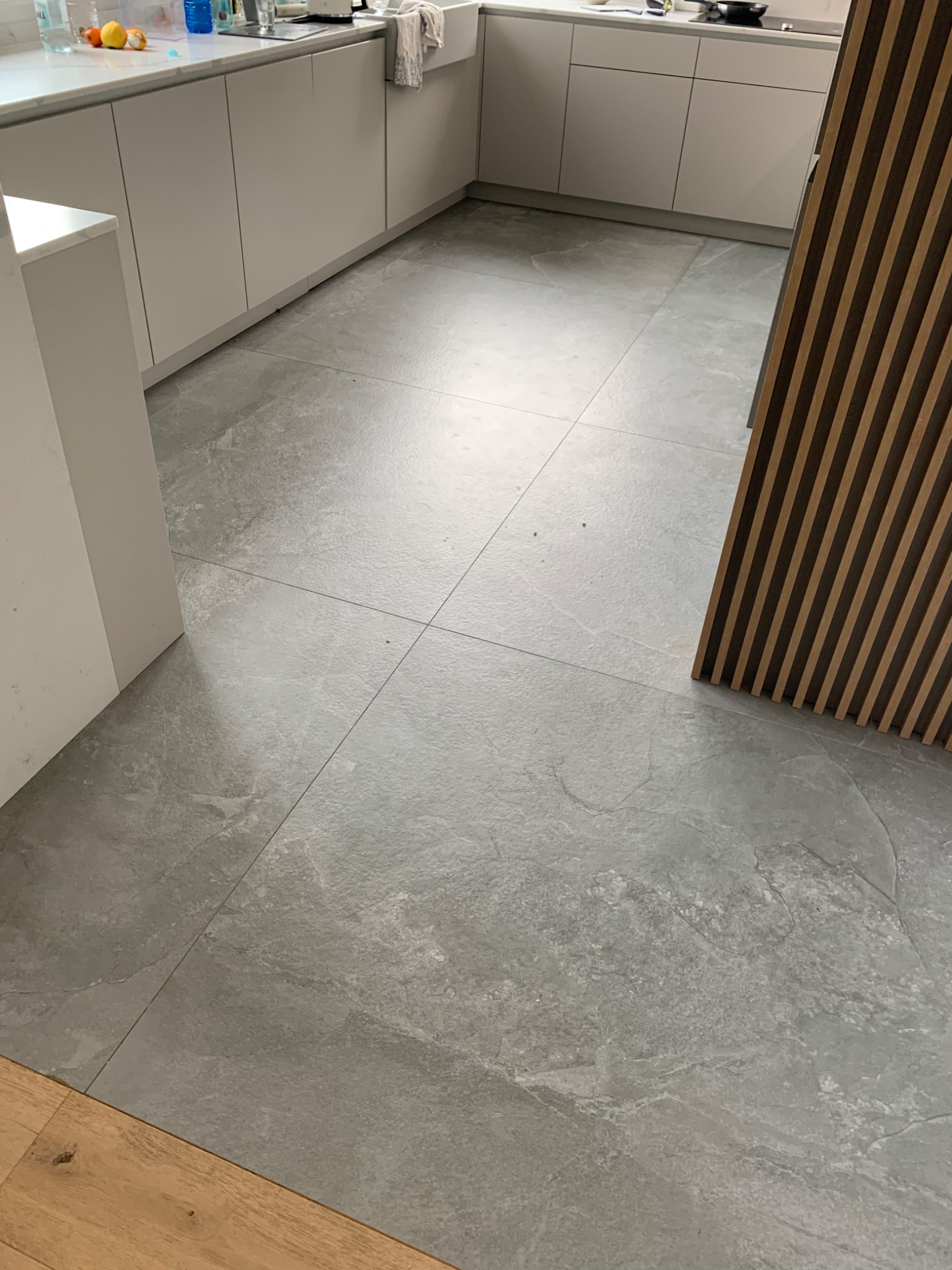 Szare płytki podłogowe w kuchni, imitujące kamień, ułożone z fugami, widoczne szafki kuchenne w tle oraz drewniana dekoracja ścienna po prawej stronie.