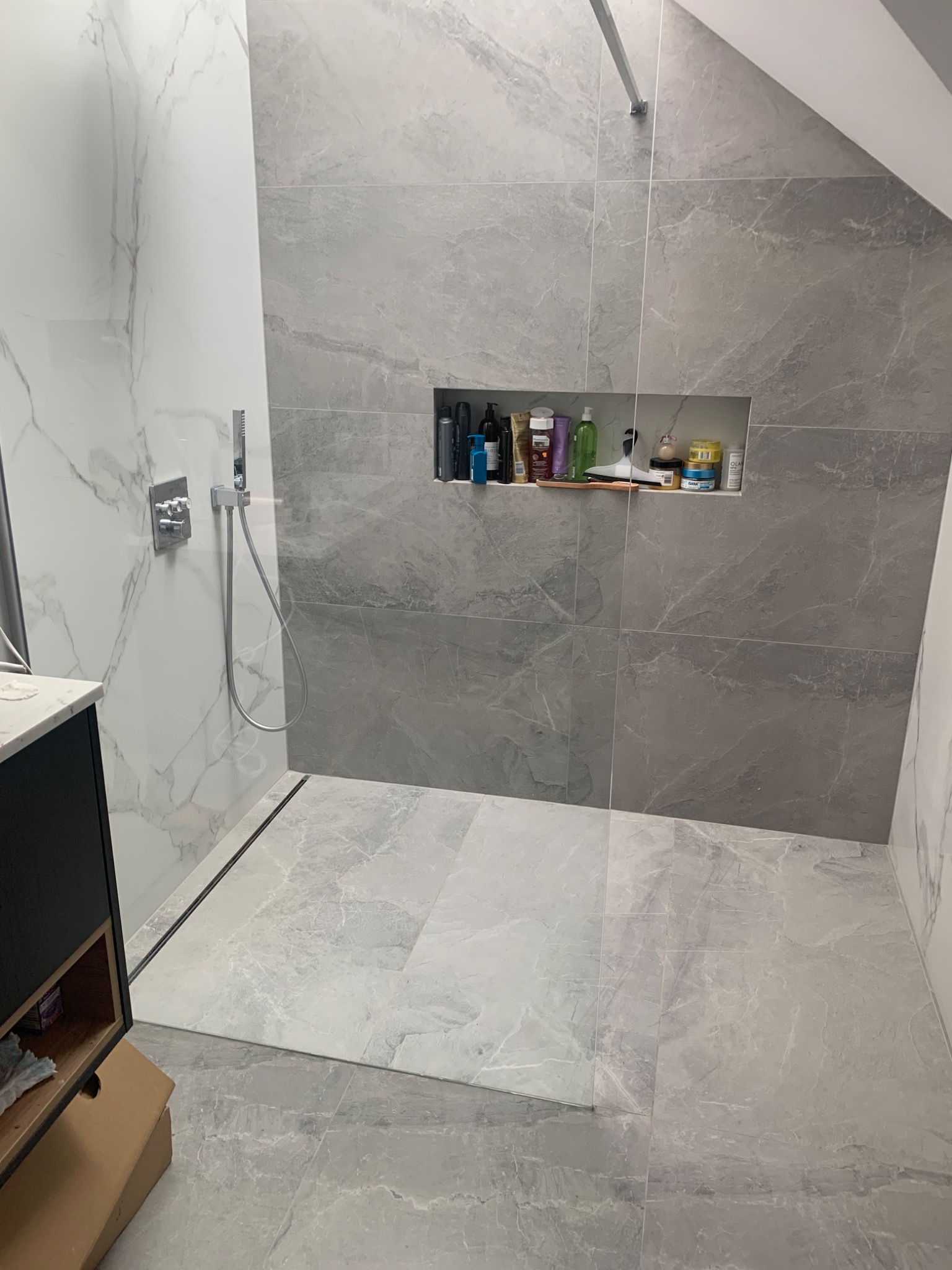 Nowoczesna kabina prysznicowa z szarymi płytkami imitującymi marmur, wnęka w ścianie wypełniona kosmetykami, chromowana armatura i odpływ liniowy.