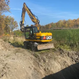 Żółta koparka JCB na skraju wykopu w słoneczny dzień, widoczne drzewa w tle.