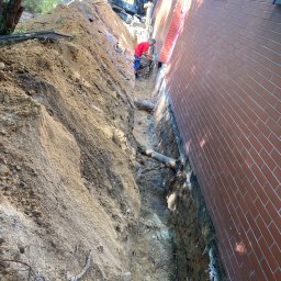 KKK Bruk - Wąski wykop wzdłuż ceglanego budynku, widoczny pracownik w czerwonej koszulce i minikoparka w tle.