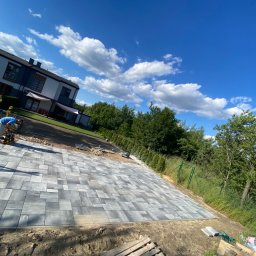 KKK Bruk - Pracownik zagęszcza wibroprasą nowo ułożoną nawierzchnię z szarej kostki brukowej o zróżnicowanych kształtach, na tle domu jednorodzinnego i błękitnego nieba z chmurami.