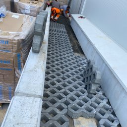 KKK Bruk - Montaż ażurowej kostki brukowej na podsypce przy budynku przemysłowym, dwóch pracowników w odzieży roboczej układa kolejne elementy, palety z kostką i materiałami budowlanymi w tle.