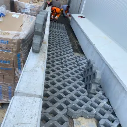 Montaż ażurowej kostki brukowej na podsypce przy budynku przemysłowym, dwóch pracowników w odzieży roboczej układa kolejne elementy, palety z kostką i materiałami budowlanymi w tle.