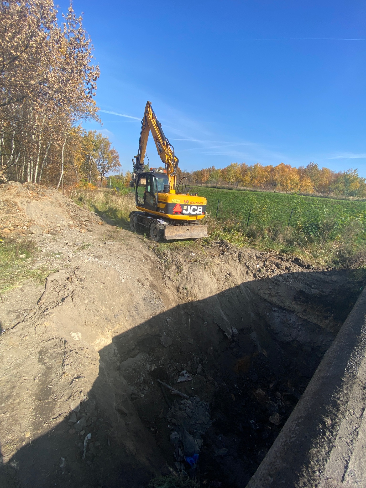 Żółta koparka JCB na skraju wykopu w ziemi, jesienne drzewa w tle pod błękitnym niebem.