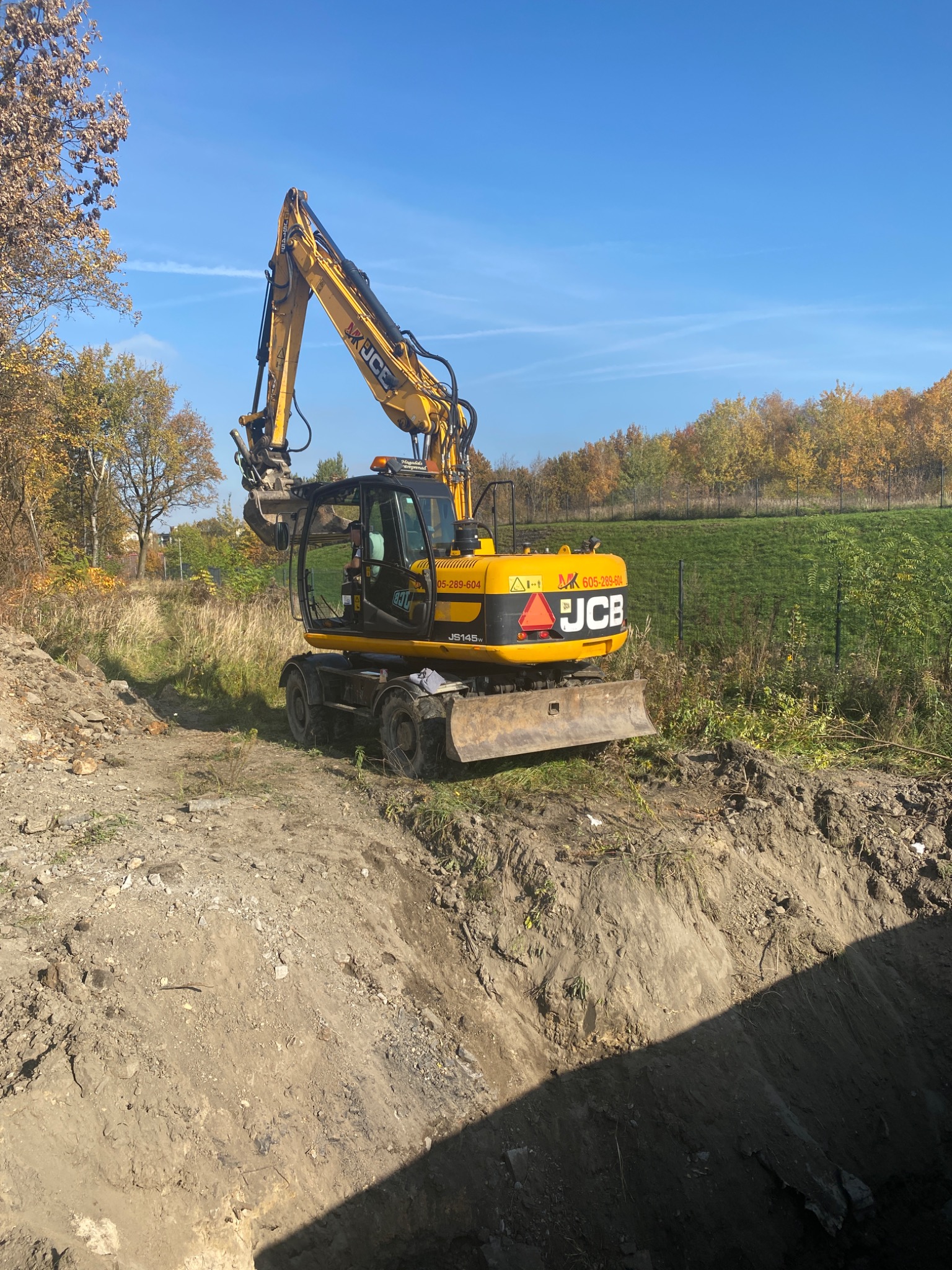 Żółta koparka JCB na skraju wykopu w słoneczny dzień, widoczne drzewa w tle.