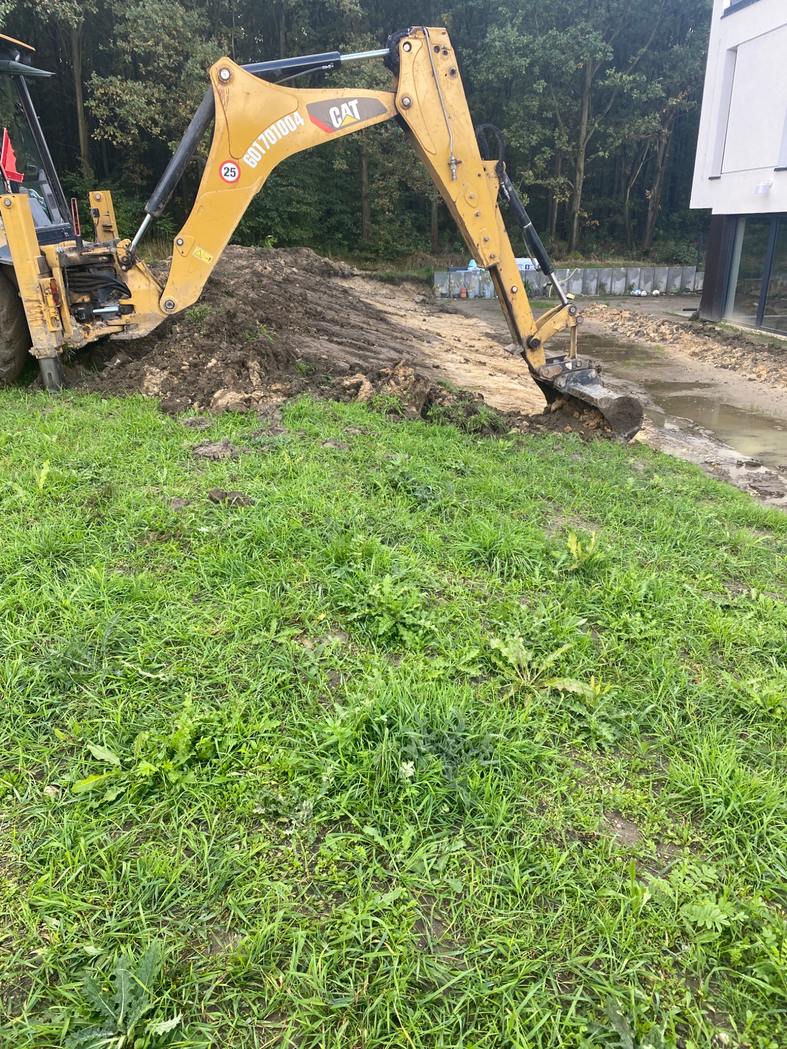 Żółta koparko-ładowarka CAT wykonuje wykop w ziemi, widoczny świeży nasyp ziemi, w tle budynek i drzewa.