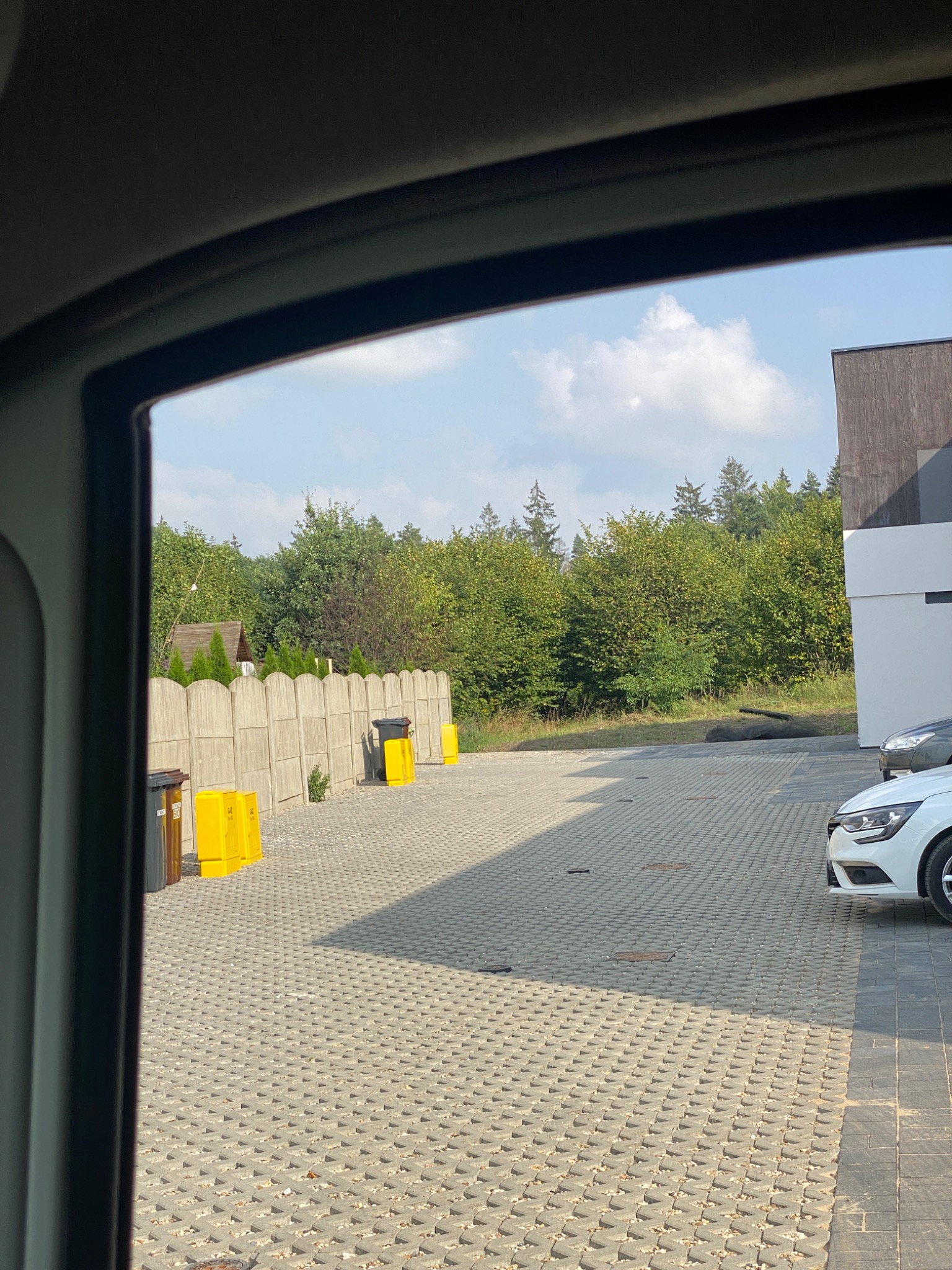 Widok na ułożoną kostkę brukową z perspektywy wnętrza pojazdu, widoczny fragment białego samochodu, betonowe ogrodzenie, drzewa oraz żółte pojemniki na odpady.