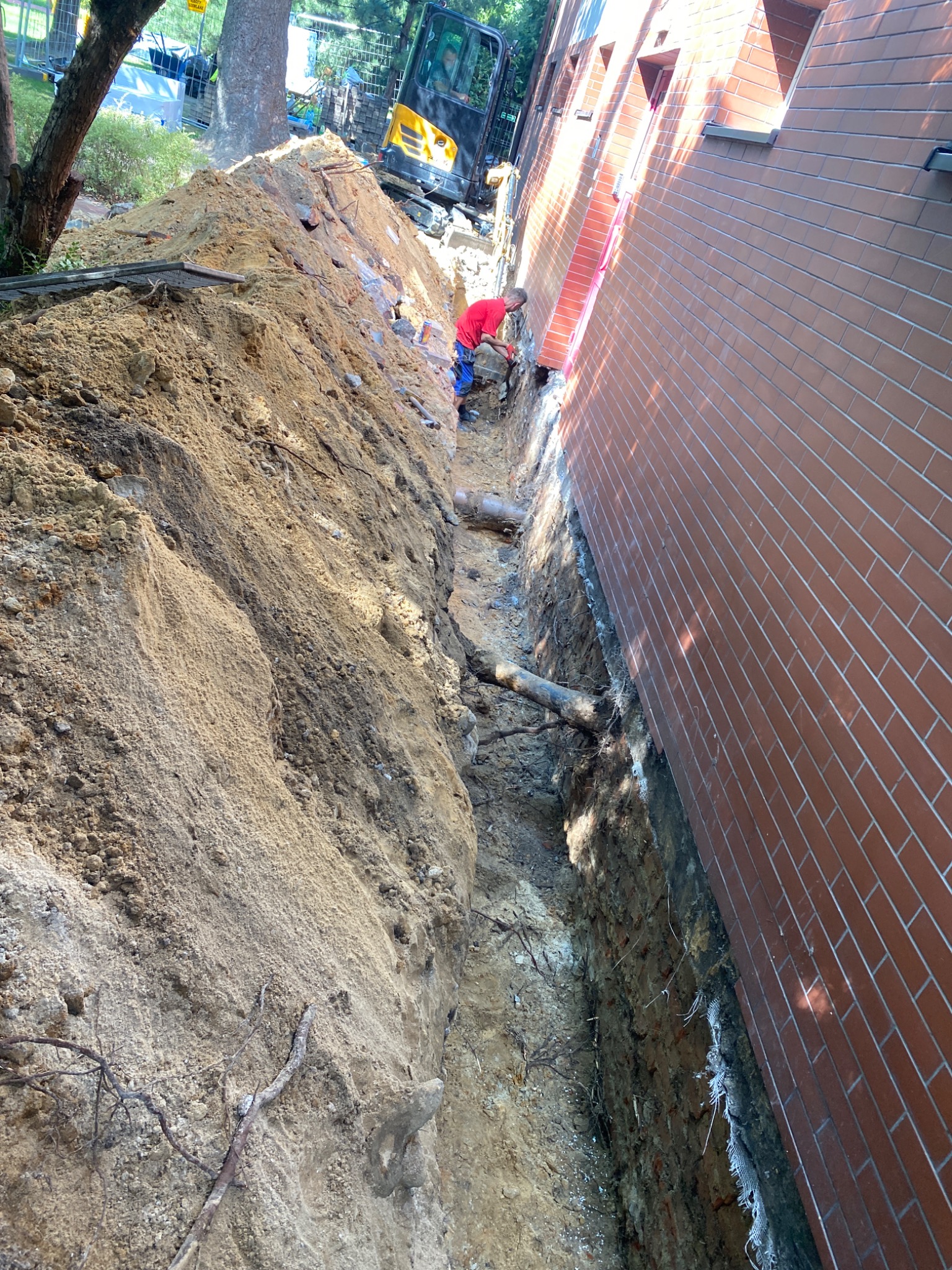 Wąski wykop wzdłuż ceglanego budynku, widoczny pracownik w czerwonej koszulce i minikoparka w tle.