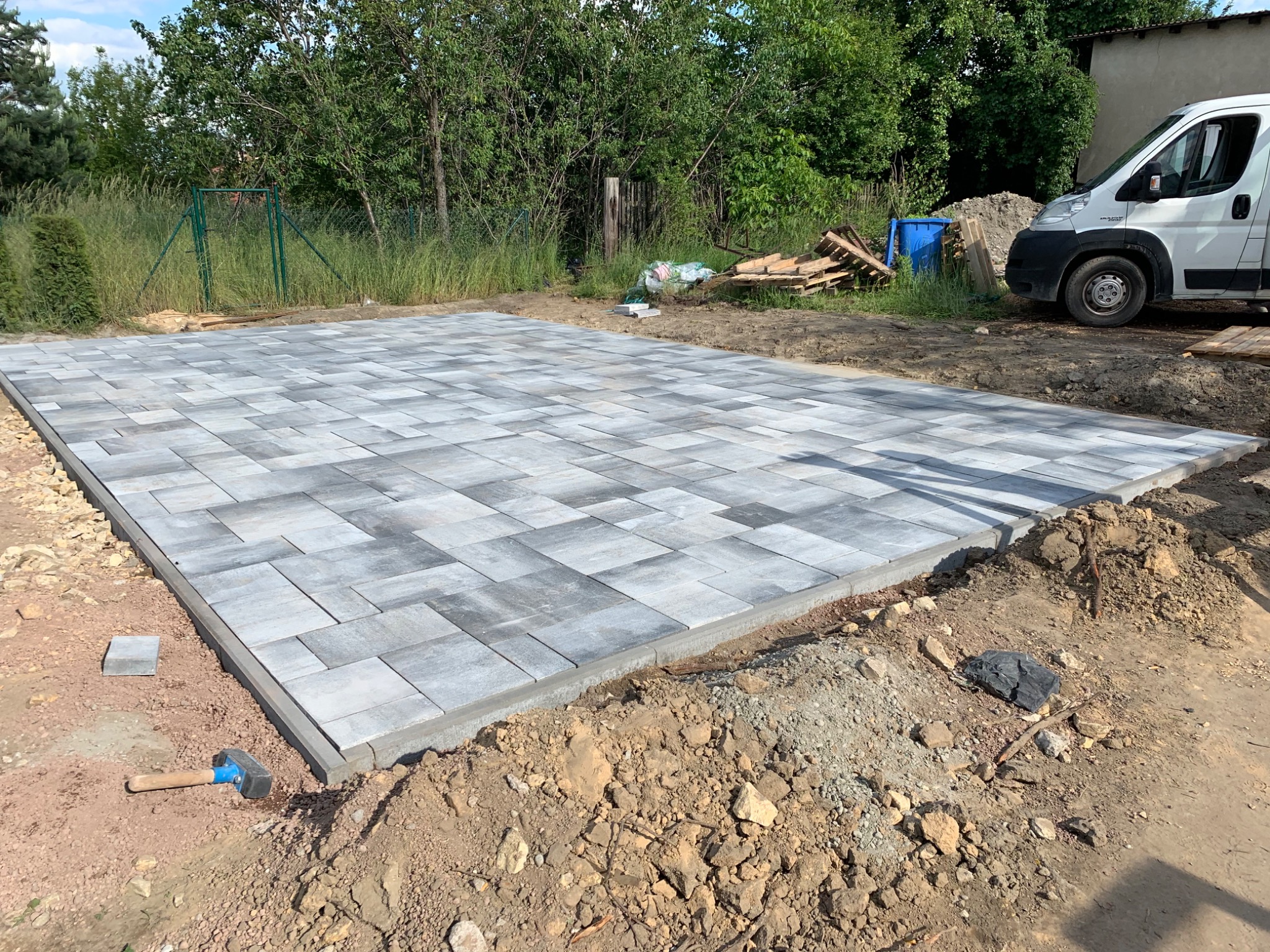 Ułożona szara kostka brukowa na przygotowanym podłożu, widoczny młotek brukarski, krawężniki i zaparkowany biały samochód dostawczy w tle.