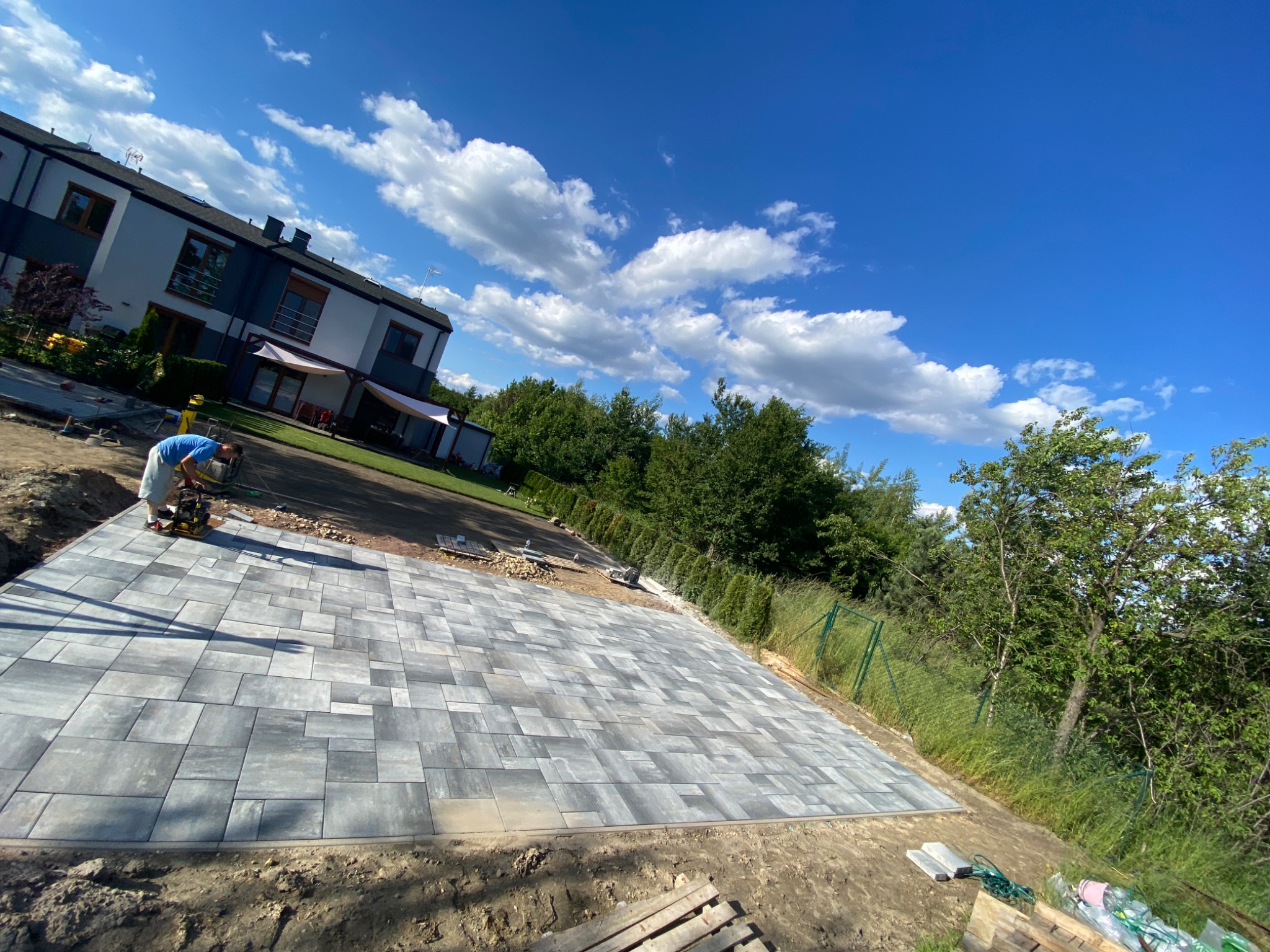 Pracownik zagęszcza wibroprasą nowo ułożoną nawierzchnię z szarej kostki brukowej o zróżnicowanych kształtach, na tle domu jednorodzinnego i błękitnego nieba z chmurami.