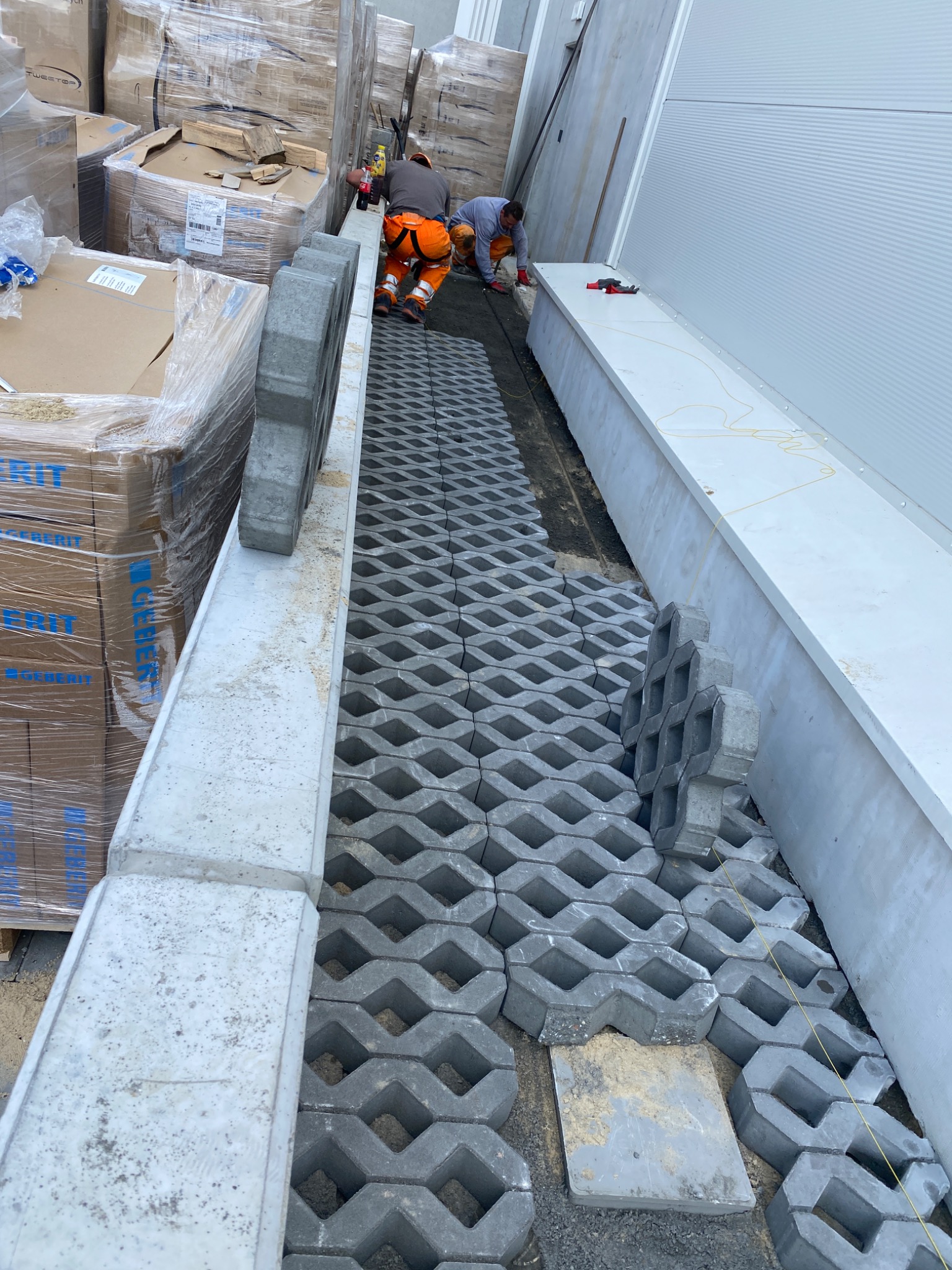 Montaż ażurowej kostki brukowej na podsypce przy budynku przemysłowym, dwóch pracowników w odzieży roboczej układa kolejne elementy, palety z kostką i materiałami budowlanymi w tle.
