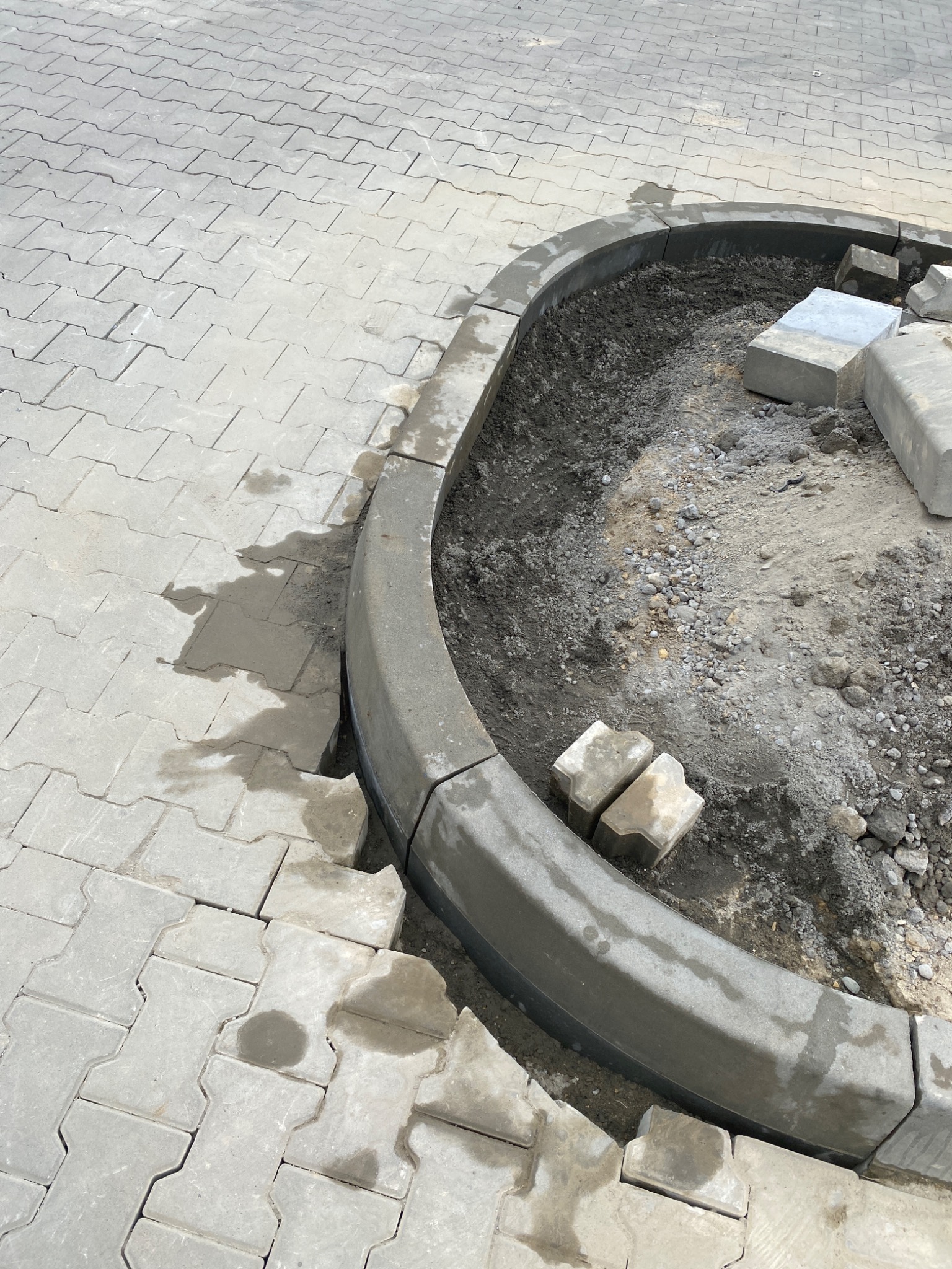 Niedokończona obudowa klombu z betonowych obrzeży i kostki brukowej, widoczne przygotowane podłoże i zapas kostki.