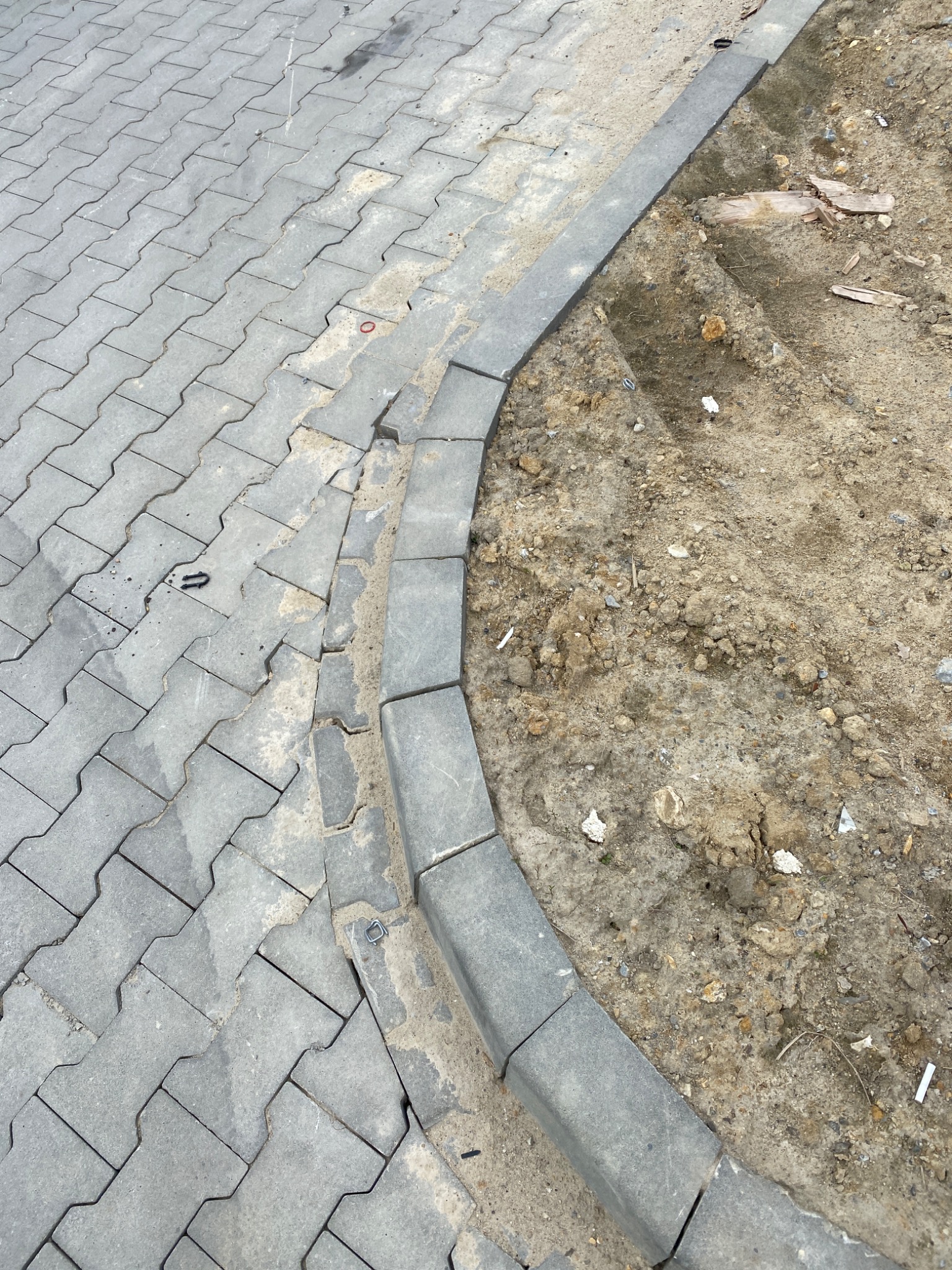 Świeżo ułożona szara kostka brukowa typu 'psi kość' z obrzeżem oddzielającym ją od ziemi z resztkami budowlanymi, widoczne niedociągnięcia przy obrzeżu.