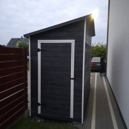 GuDREW - Drewniana, pomalowana na szaro budka narzędziowa z białym obramowaniem drzwi, ustawiona na trawniku obok ogrodzenia i chodnika z kostki.