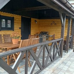 GuDREW - Zadaszony taras z drewnianą konstrukcją, wyposażony w okrągły stół z krzesłami, grill i dekoracje w postaci podków na ścianie.