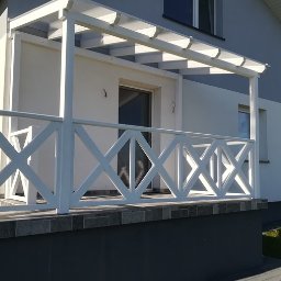 GuDREW - Nowoczesny taras z białą balustradą w kształcie krzyży i zadaszeniem pergolowym, przylegający do budynku z szarą elewacją i widocznym logo firmy w prawym dolnym rogu.