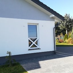 GuDREW - Elewacja domu z oknem zabezpieczonym białym krzyżem, widok z kostki brukowej na podwórzu.