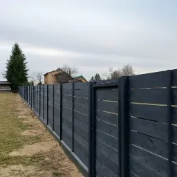 Długa, nowoczesna, pozioma, drewniana, pomalowana na ciemnoszaro ogrodzenie z metalowymi słupkami i furtką, oddzielająca posesję od otoczenia, widoczne domy w tle.