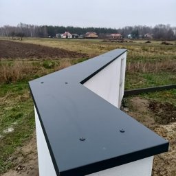 GuDREW - Wykończony fragment ogrodzenia z białego betonu z ciemnoszarym metalowym daszkiem, widoczne wkręty mocujące, w tle pole i zabudowania.