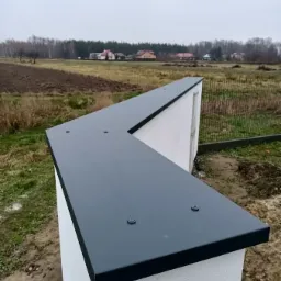 Wykończony fragment ogrodzenia z białego betonu z ciemnoszarym metalowym daszkiem, widoczne wkręty mocujące, w tle pole i zabudowania.