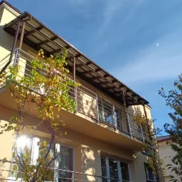 Dwupoziomowy budynek z balkonami z metalowymi balustradami i zadaszeniem, pnącza z żółtymi liśćmi na elewacji, błękitne niebo w tle.