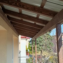 GuDREW - Zadaszenie tarasu z drewnianą konstrukcją nośną i metalowym pokryciem, widok od dołu z fragmentem elewacji budynku.