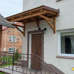 GuDREW - Drewniany daszek nad wejściem do domu z brązowymi drzwiami, oświetlony lampą, z widocznymi schodami i metalową barierką.