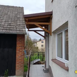 Drewniany daszek nad wejściem do domu z widoczną konstrukcją wsporczą, brązowe pokrycie, ściana budynku z fakturą, na parapecie stoi biały kubek.