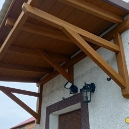 GuDREW - Drewniany daszek nad wejściem do domu, wsparty na solidnych belkach, z oświetleniem zewnętrznym, widok z dołu.