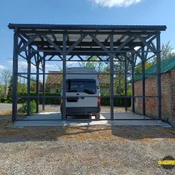 Stalowa konstrukcja z zadaszeniem, prawdopodobnie wiata garażowa, z zaparkowanym białym busem w środku, widok z zewnątrz, na tle budynków i zieleni.
