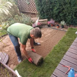 Mężczyzna w zielonej koszulce rozwijający rolkę trawy na przygotowanym podłożu z piasku, obok drewniany taras z narzędziami ogrodniczymi i butelką wody, w tle roślinność ogrodowa.