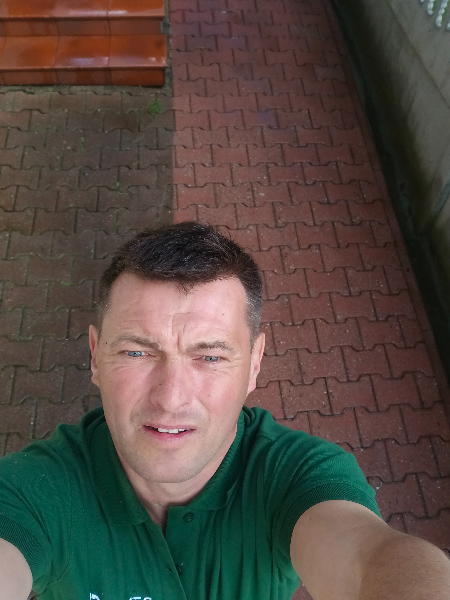 Mężczyzna w zielonej koszulce, robiący selfie na tle kostki brukowej, której część jest wyraźnie czystsza.