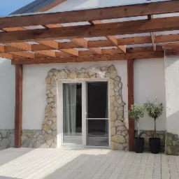 Zadaszony taras z drewnianą konstrukcją, wykończony kamieniem dekoracyjnym wokół drzwi, z drewnianą podłogą i dwoma drzewkami oliwnymi w donicach.