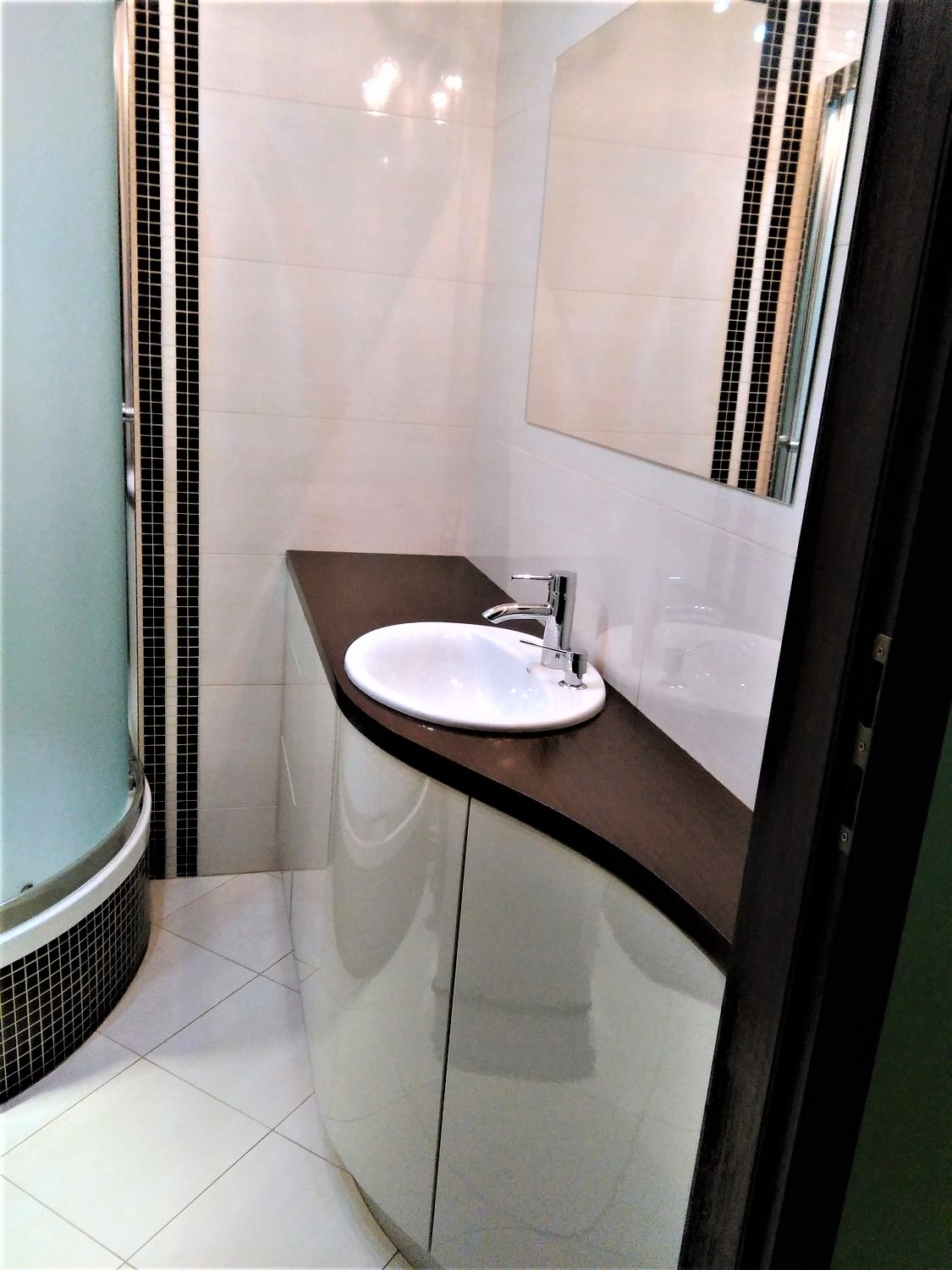 Nowoczesna łazienka z umywalką nablatową na szafce o zaokrąglonych frontach, kabina prysznicowa z mozaiką i lustro na ścianie wyłożonej białymi płytkami.