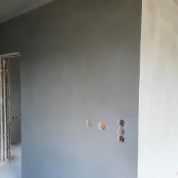 Wnętrze w trakcie prac wykończeniowych: ściany pokryte świeżym, szarym tynkiem maszynowym, widoczne puszki elektryczne i surowa ościeżnica drzwiowa.