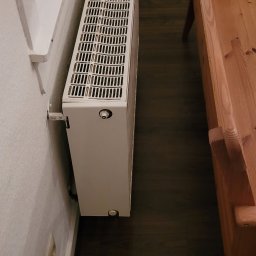 Sergeii Shystia - Biały, żebrowany grzejnik przymocowany do ściany obok okna, widoczna podłoga z ciemnego drewna i fragment drewnianego krzesła dziecięcego.