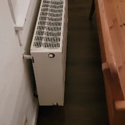 Biały, żebrowany grzejnik przymocowany do ściany obok okna, widoczna podłoga z ciemnego drewna i fragment drewnianego krzesła dziecięcego.