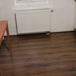 Sergeii Shystia - Wnętrze pokoju z białymi ścianami, drewnianą podłogą, grzejnikiem pod oknem i fragmentem stołu po lewej stronie. W oknie widoczne odbicie osoby robiącej zdjęcie.