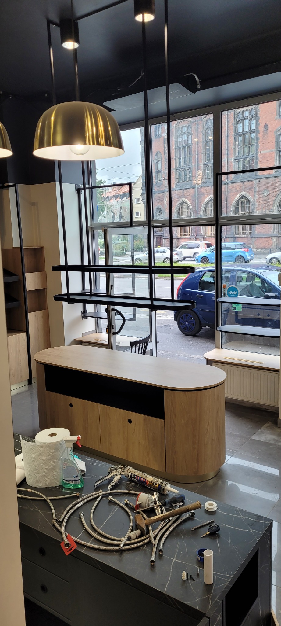 Wnętrze sklepu w trakcie remontu, widoczne narzędzia i materiały budowlane na blacie, w tle okno z widokiem na budynek z czerwonej cegły.